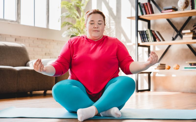 Plump obese caucasian fat woman meditating in sporty outfit at home. Fat woman sitting and relaxing